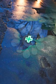 green cross of drugstore reflected in rain puddle in dark urban evening