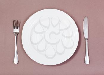 top view of empty white plate with fork and knife on brown background