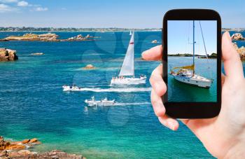travel concept - tourist taking photo of yacht near Brittany coastline on mobile gadget, France