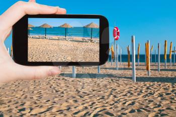 travel concept - tourist taking photo of straw beach umbrellas on ocean coast, Portugal, Algarve on mobile gadget
