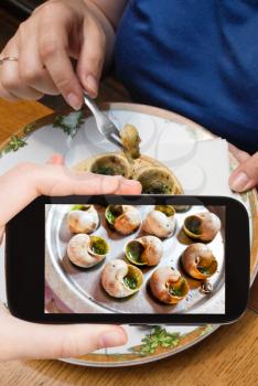 photographing food concept - tourist takes picture of hot plate of escargot shells, with special tongs and fork on smartphone,
