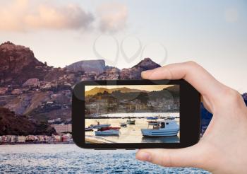 travel concept - tourist takes picture of boats at sunset, near town Taormina, Sicily, Italy on smartphone