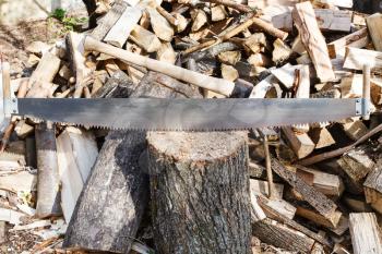 two-handled saw and cleaver in chopping block, pile of firewood on rustic courtyard