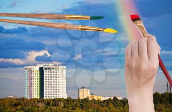 nature concept - hand with paintbrush paints rainbow in blue clouds over urban houses