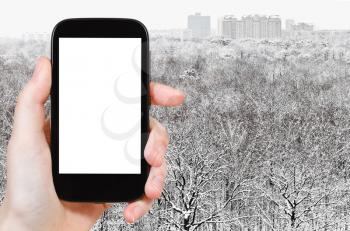 travel concept - hand holds smartphone with cut out screen and snowy forest and city on background