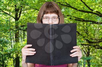 girl with glasses reads big book with blank cover and green summer forest on background