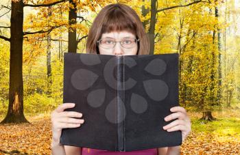 girl with glasses reads big book with blank cover and yellow autumn woods on background