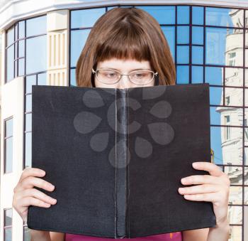 girl with glasses reads big book with blank cover and modern apartment house on background