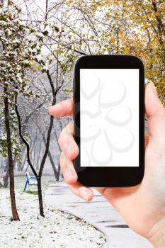 season concept - hand holds smartphone with cut out screen and first snowfall in city park on background