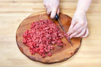 woman finely dices meat on wooden cutting board on table