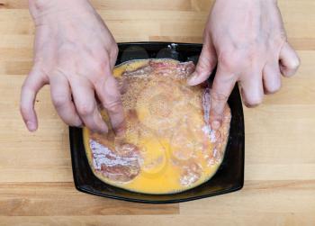top view of cook hands bread a raw veal schnitzel in flour and eggs in bowl on wooden table