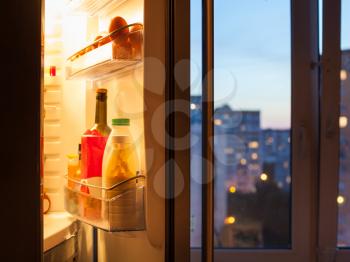 Open door of refrigerator with food and view of city through home window in evening