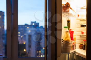 Open door of refrigerator with meal and view of city through window in evening