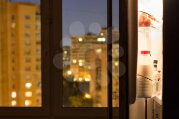Open door of home fridge and urban view through window glass in night