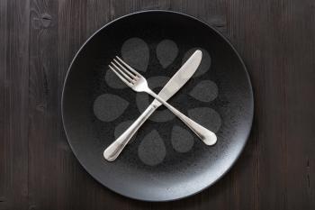 food concept - top view of black plate with crossing knife, spoon on dark brown table