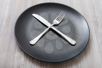 food concept - black plate with crossing knife, spoon on gray brown table