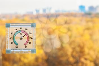 outdoor thermometer on home window and blurred yellow city park on background in sunny warm autumn day