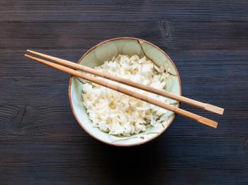 top view of chopsticks above boiled rice in cup on dark brown table with copyspace