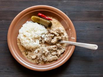 Russian cuisine dish - eating of Beef Stroganoff (Beef Stroganov, Befstroganov) pieces of stewed meat in sour cream with boiled rice on brown plate on dark table