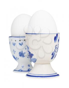 side view of two white boiled eggs in ceramic egg cups isolated on white background, the egg with a blunt end up on foreground