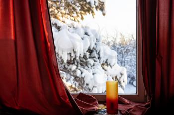 electric night lamp on window sill and red curtain in cottage and view of snow-covered pine tree at backyard in cold winter evening (focus on the curtain)