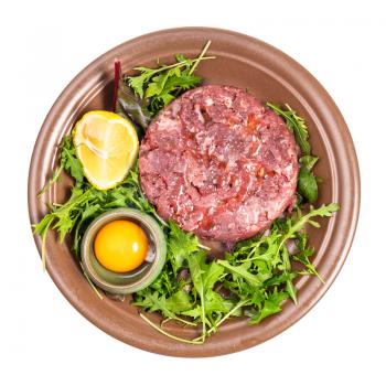 top view of portion of Steak tartare (raw minced beef meat and raw yolk in bowl on fresh greens) on brown plate isolated on white background