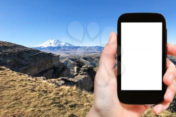 travel concept - tourist photographs of cliff of Bermamyt Plateau and view of Mount Elbrus in North Caucasus mountains of Russia on smartphone with cut out screen with blank place for advertising