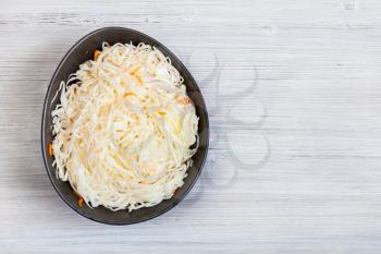 top view of Russian sauerkraut (sour cabbage pickled with carrots and served as salad) in black bowl on gray wooden background with copyspace