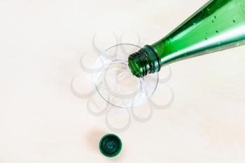 top view of pouring sparkling mineral water from green plastic bottle in glass on light brown table close up