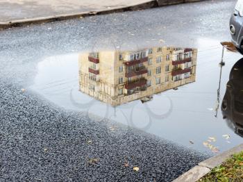 rain puddle with reflection of municipal urban house on asphalt road in city on autumn day