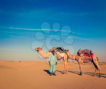 Vintage retro hipster style travel image of Rajasthan travel background - Indian cameleer (camel driver) with camels in dunes of Thar desert. Jaisalmer, Rajasthan, India