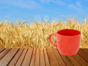 Red tea mug on wooden surface against of golden wheat ears and blue sky.