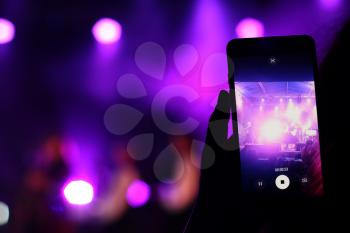 Shooting festival concert on smartphone with place for text. Blurred music stage bokeh background for design. Fans takes picture of scene on phone