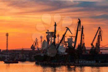Varna port at sunset under dramatic cloudy sky. Black silhouettes of cranes, port buildings and ships