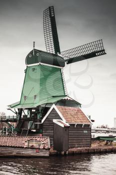 Windmill under cloudy sky on Zaan river coast, Zaanse Schans town, popular tourist attractions of the Netherlands. Suburb of Amsterdam