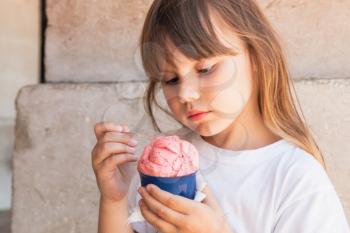Little blond European girl eats pink ice cream, close up outdoor portrait 