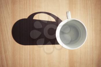 Empty white coffee cup on wooden table background with strong dark shadow from the sunlight. Retro photo filter effect