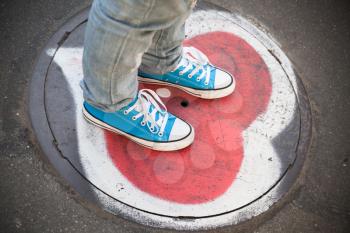 Blue sneakers, teenager feet stand on urban sewer manhole with heart sigh