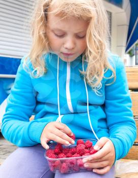 Little blond girl takes raspberry from full plastic box. Outdoor portrait