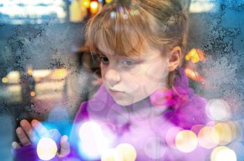Little blond girl looks through frozen shop window glass with colorful blurred lights