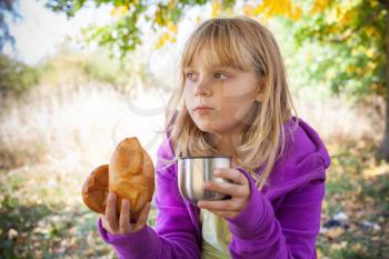 Little blond girl in autumn park eats small pies with tea