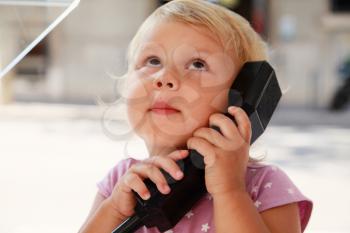 Outdoor portrait of surprised little Caucasian blond girl talking on the street phone