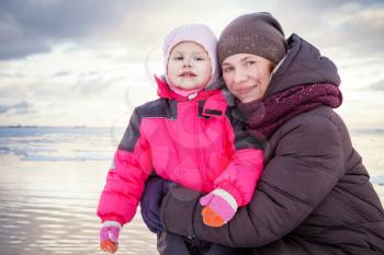Caucasian family outdoor portrait on the winter sea coast, young mother hugs her little baby girl in pink jacket