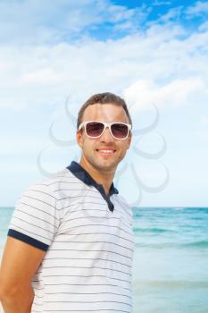 Outdoor portrait of young positive Caucasian man standing with white sunglasses on summer sea coast