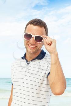 Outdoor portrait of young smiling Caucasian man standing with white sunglasses on summer ocean coast