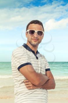 Outdoor portrait of young smiling Caucasian man standing with white sunglasses on the summer sea coast. Vintage toned photo with style filter effect
