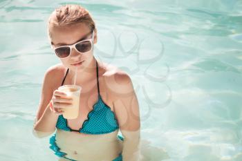 Little blond girl drinks cocktail in swimming pool, toned photo filter effect
