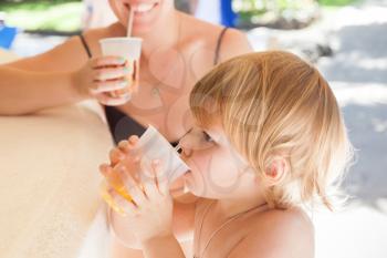 Blond Caucasian baby girl with young mother drink orange juice in an open resort bar