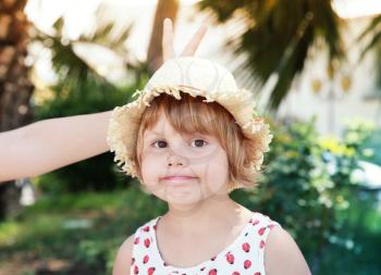 Cute Caucasian little girl in straw hat, other person makes horns with fingers