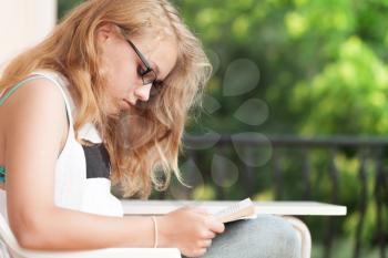 Blonde Caucasian teenage girl sits on the balcony and read a book